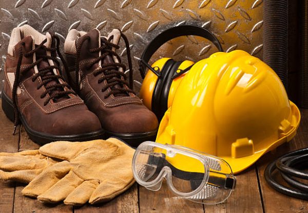 Personal Protective Workwear Shoot on Work Location. The Safety items are placed on rustic wood and includes a Yellow Hard Hat, Gloves, Steel Toe Shoes, Ear Muff and Goggles. A silver diamondplate in the background. Predominant colors are yellow and brown